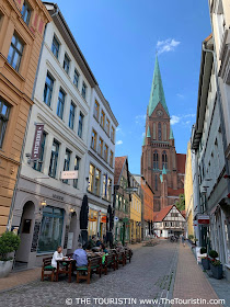 People sitting in a cafe on a cobblestone lane that leads to a large red-brick cathedral.