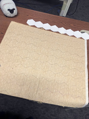 A fat quarter of yellow fabric with faint white dots and dashes, laid out wrong-side-up on a medium-brown wood table, with faint pencil lines marking zigzags that describe rows of hexagons over the whole piece. A white paper template of nine hexagons in a row is just above the top edge of the fabric. A white stuffed sheep with a black face looks on from the upper left corner of the fabric.