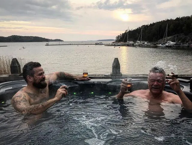 Two guys smiling big smoking cigars and a hot tub on the deck of a boat with beautiful bay in backgrojnd