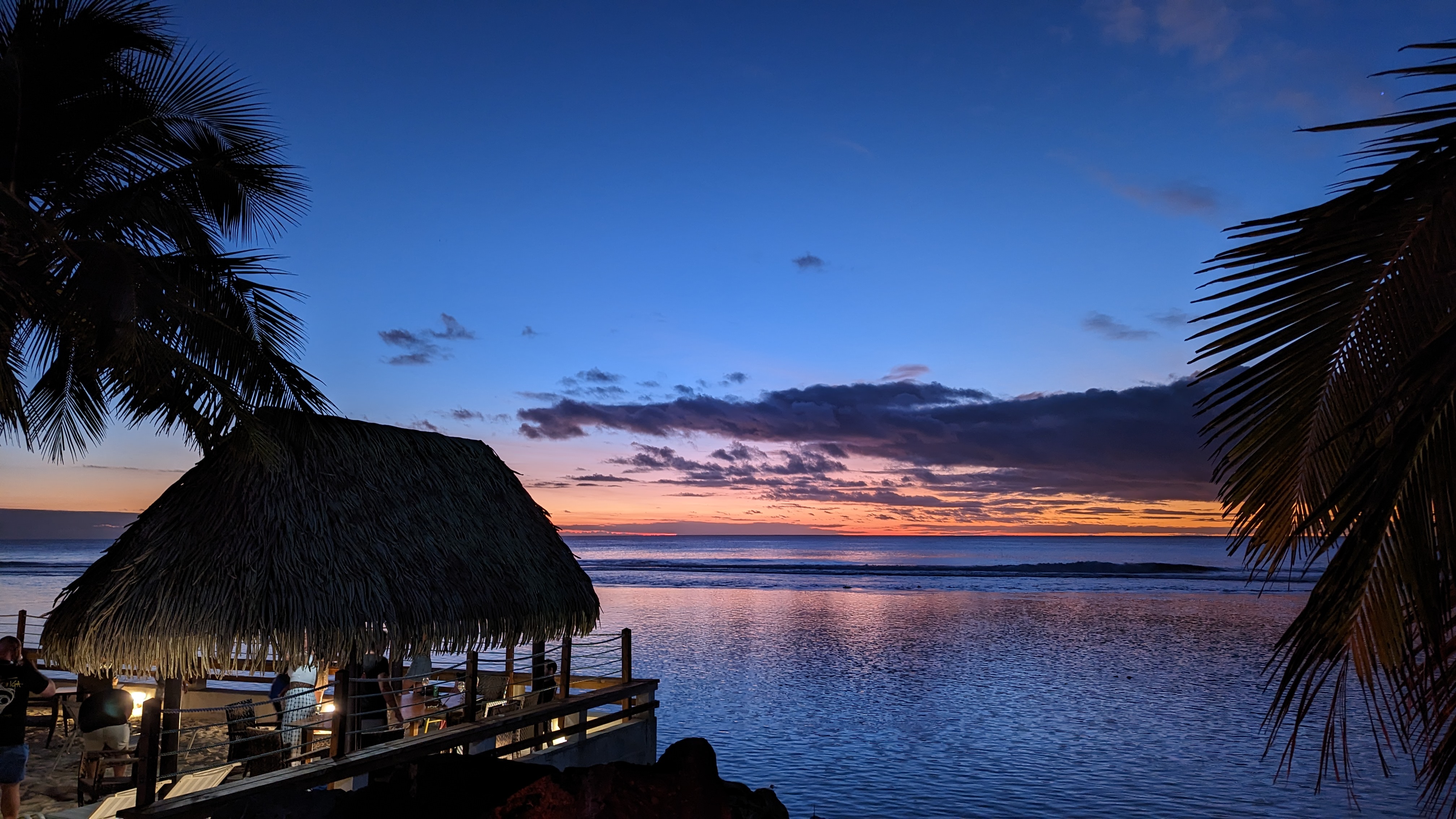 Blues of the darkening sky against a sliver of orange sun, all reflected in the calm ocean. In the foreground is a resort thatched bar and palm trees.