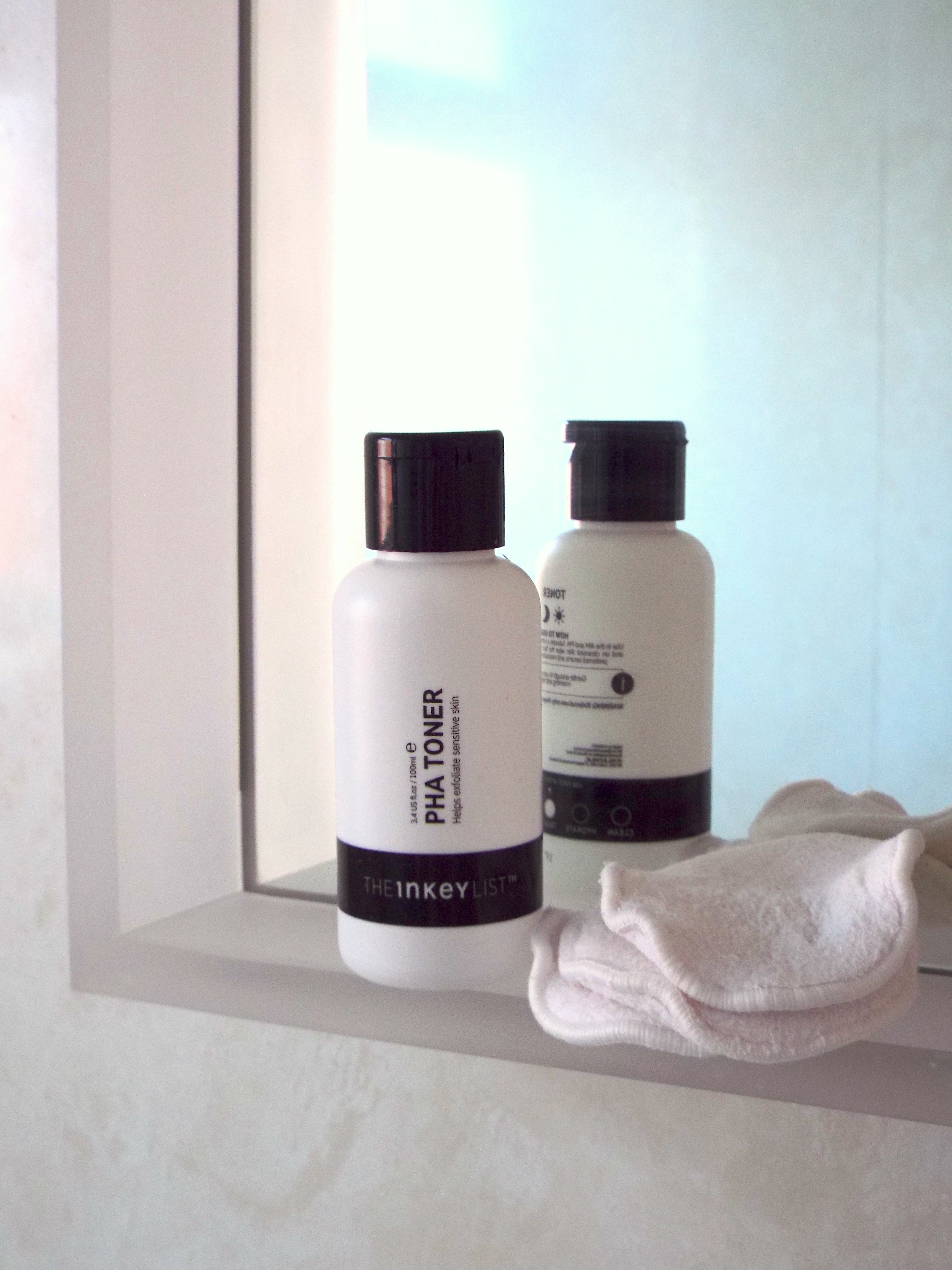 White opaque black and white The Inkey List PHA Toner bottle sat on bathroom mirror-side, adjacent to pile of reusable cotton pads.