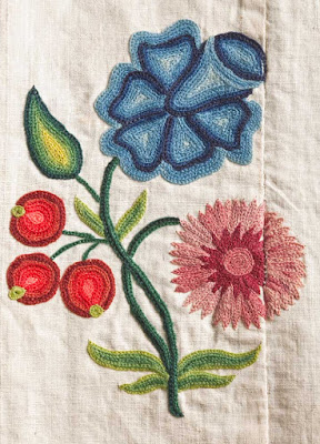 A chain-stitched sprig of blue flower, pink carnation, three red cherries, and a few green leaves. A seam runs through the right edge of the carnation.