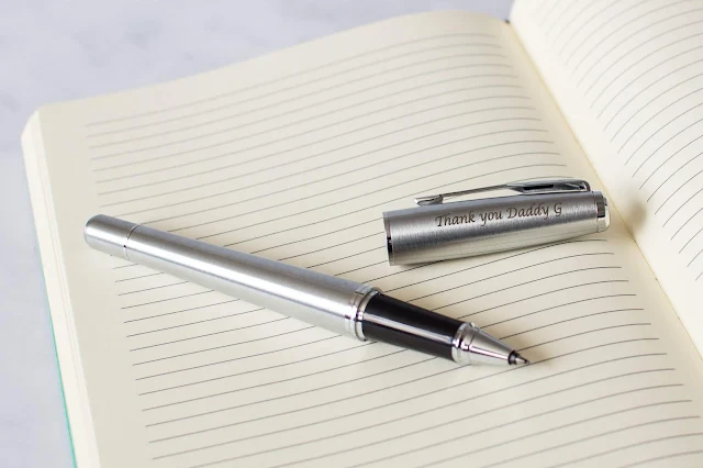 A silver coloured roller ball pen by Parker at Pen Heaven with etching on the lid saying "Thank you Daddy G" on an open lined notepad