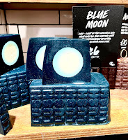 A photo of some slices of a rectangular blue soap all containing a white moon in the middle with a black card next to it that says blue moon soap in white font on a large light brown rectangular shelf on a bright background