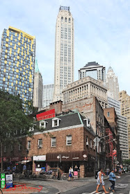 Back on dry land in lower Manhattan, a low-rise 19th century brick building on Coenties Alley with Art Deco skyscrapers, including 20 Exchange Place, of the financial district rising in the background. Travel photography by Kent Johnson.