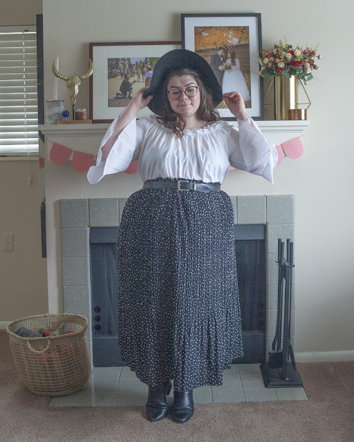 An outfit consisting of a black wide brim hat, white off the shoulder blouse tucked into a black and white dotted and pleated maxi skirt and black chelsea boots.