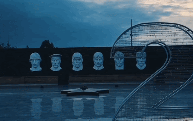 A solemn war memorial featuring busts of soldiers in uniform, set against a dark wall at dusk in Ganja, Azerbaijan, with a large decorative number 2 in the foreground.