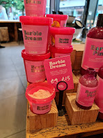 A wide shot of some tall cylindrical pink plastic tubs filled with pink and white sugar scrub with pink lids stacked on top of each other all with rectangular pink labels that say barbie dream sugar scrub in white font on a large light brown shelf with a pink rectanguoar label next to it that says barbie dream sugar scrub in white font on a bright background