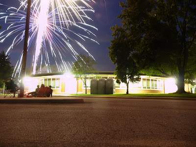 A family is sitting outside of a round school at evening. A firework is exploding above the school.