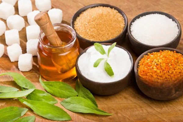 An assortment of sugar alternatives including honey, raw sugar, stevia leaves, and granulated sweeteners displayed on a wooden surface.