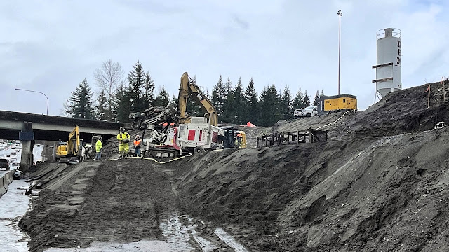 Crews build support structures near the I-90/SR 18 interchange. Crew members and machinery work atop a slope that leads to State Route 18 beneath an I-90 overpass.