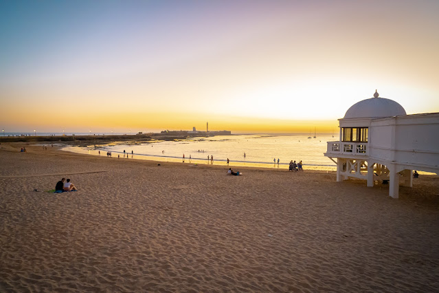 Arena de playa en el primer plano con el agua del mar de color dorado por los rayos del sol al atardecer, además de la estructura de un antiguo balneario.