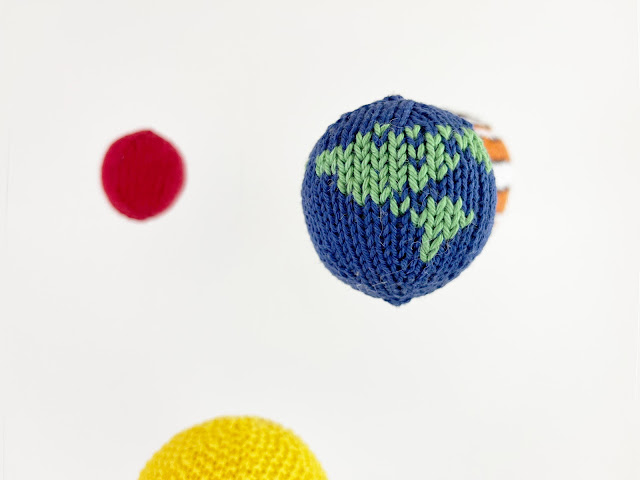 close up of knitted planet earth with sun, jupiter and mars around it as part of the solar system space mobile hanging in front of a white background