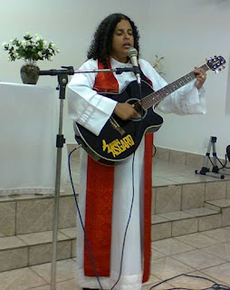 Rev. Borges standing on a platform, she is wearing a pastor's robe with a red stole, and playing a guitar. 