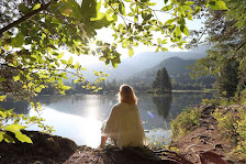 A woman sits on the shore of a lake among trees.