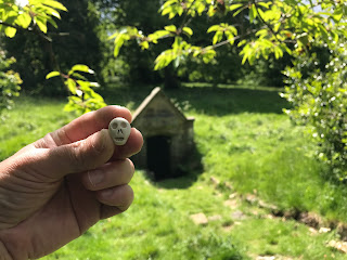 A photo of a small ceramic skull (Skulferatu 77) being held up in front of the Oyly Well.  Photograph by Kevin Nosferatu for the Skulferatu Project.