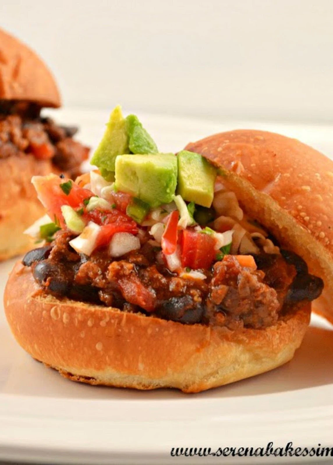 A hamburger bun on a white plate topped with Mexican Sloppy Joe filling, pico de gallo, and avocado.