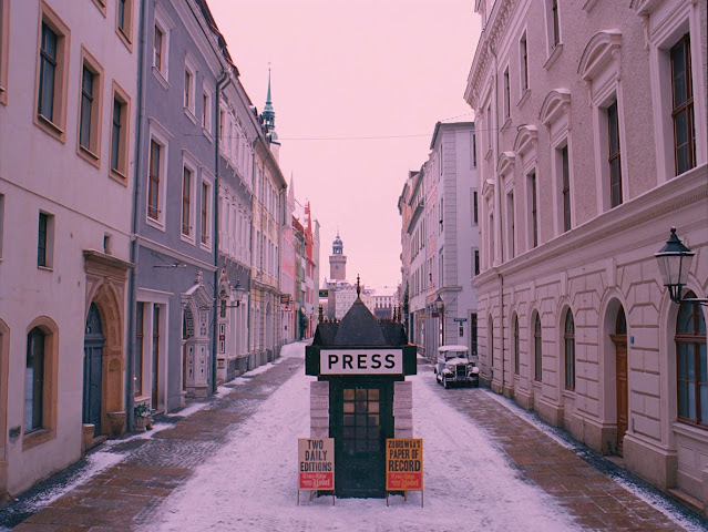 Cena de "O Grande Hotel Budapeste". Mostra uma rua de uma cidade europeia, há casas em ambos os lados e neve no chão. No meio da rua há uma cabine de jornaleiro antiga.