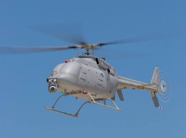 Un helicóptero no tripulado de color gris, que carece de cabina o de cualquier tipo de ventana, se muestra flotando en un cielo azul y claro. Lleva un sensor que apunta hacia abajo bajo su nariz.