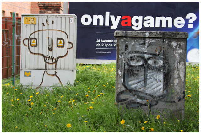 Caras pintadas con spray en dos cajas de electricidad, y de fondo una pancarta publicitaria que deja ver un spot diciendo " only a game?"