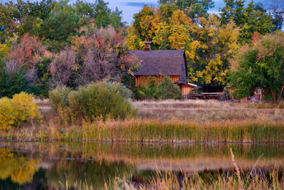 An old barn nestled among trees with changing colored leaves and reflected in the pond