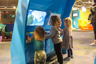 three small children play with an interactive exhibit; two screens are visible on a colorful blue curved background that extends above their heads, at the Kirby Science Discovery Center in Sioux Falls, South Dakota