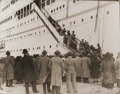A large crowd of people stand on the dock, watching passengers descend the stairs from an ocean liner. Black and white.