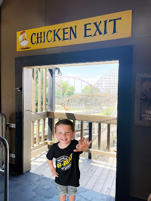 A white boy with brown hair in a black shirt and camo pants stands in a doorway with a rollercoaster in the background. Above the doorway is a sign that says "Chicken Exit."