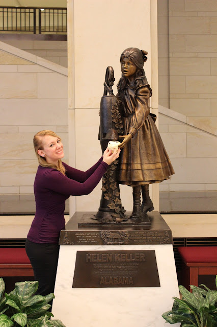 A blonde young woman of about 5'4" wearing a long sleeved purple shirt and black pants stands sideways with her head turned to smile at the camera.  She has arms extended up bronze statue of Helen Keller as young girl water pump that helped Annie Sullivan teach her ASL.  In the blonde's hands is small plush yellow star face used tactile tool for shows planetarium.  The statue extends about three feet above the woman.  It sits white pedestal plaque which states representing state Alabama.  All text is also printed and in Braille.  The room in which the picture was taken has stone walls and was bright due skylights overhead that are not visible in the picture.