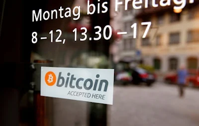 A storefront window displaying business hours in German, with a sticker indicating "Bitcoin Accepted Here" reflecting the growing adoption of cryptocurrency in everyday transactions.