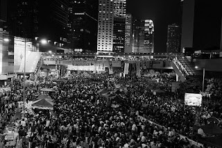 Black and white photograph of a large crowd of people, possibly numbering thousands, some carrying placards, on the streets of a city at night. There are high-rise buildings and neon lights in the background.