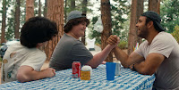 3 young men sit at an outdoor picnic table, 2 of them arm wrestling