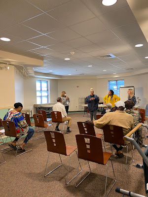 Participants are sitting in seats at the first Talk With A Doc at West End Branch Library. They are listening to two doctors, standing at the front of the room.