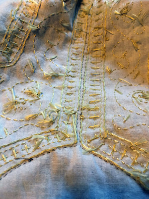 White linen showing the backside of gold embroidery, with lots of knots and frayed thread ends, and a tiny line of tan stitching outlining the just-visible curve of a round keyhole neckline at the bottom of the photo, and continuing down both sides of the neck slit.