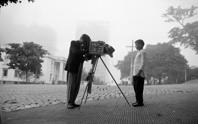 Um homem fotografa um menino com uma câmera antiga apoiada em um tripé. O homem está posicionado atrás da câmera, e o menino está parado com o corpo reto de frente para a câmera. O fundo da fotografia é composto pela vista de uma cidade, com uma casa e uma árvore no canto esquerdo, e mais árvores na outra extremidade.