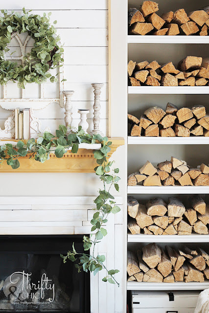 living room with white couch, large window, stacked wood, and shiplap mantel