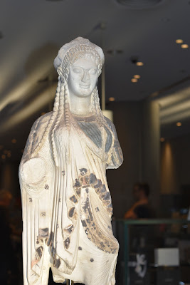 Primeira escultura mostrada é a Kore 675. Está em pé vista de frente com uma manta drapeada e com detalhes no vestido