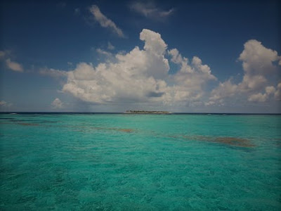 Une île se dessine à l'horizon. L'eau est turquoise et semble peu profonde. Quelques nuages blanc planent dans notre direction.