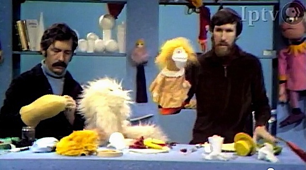 Don Sahlin, with think black hair and mustache, and Jim Henson, with brown hair and long scruffy beard, creating puppets from various materials on table before them