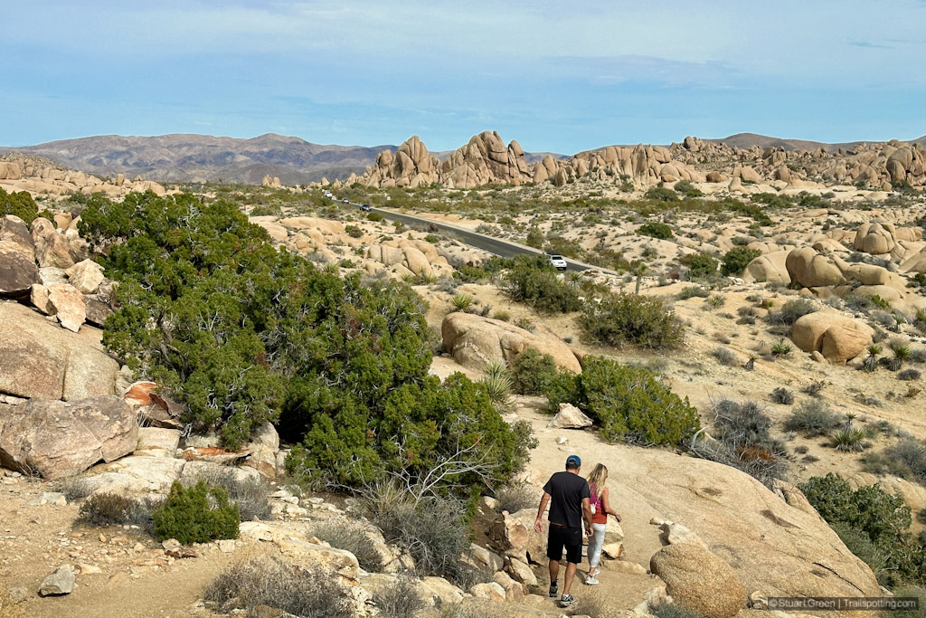 Two hikers on a dusty stepped trail, heading for Jumbo Rocks. A highway intersects the photograph.