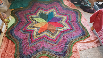 Photo of a large, colourful, star shaped blanket lying on some towels and foam mats. There is some clutter and mess around it.