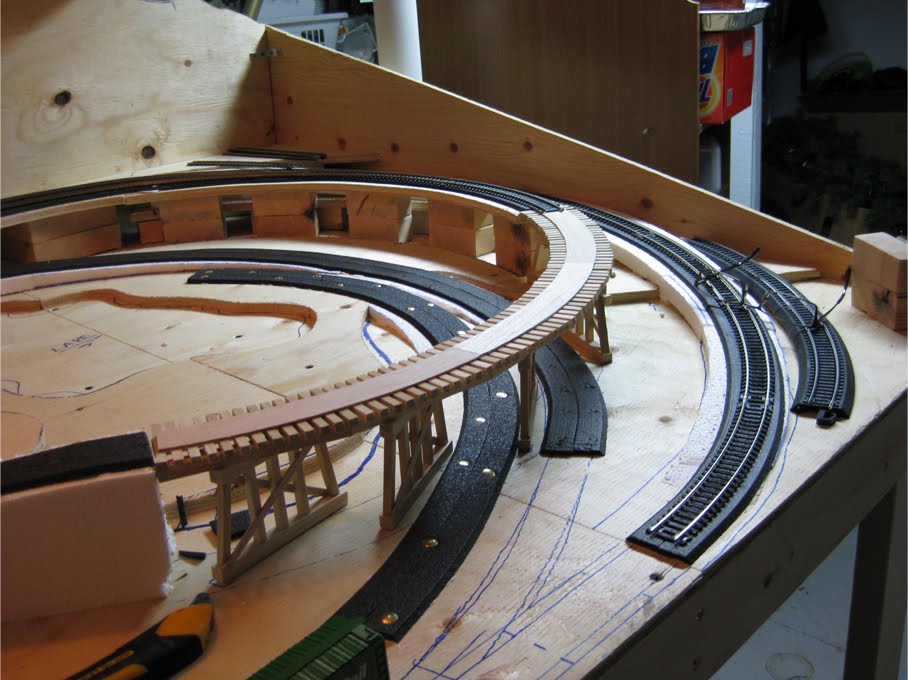 An unpainted wooden train trestle made of wooden dowels and basswood set on the benchwork