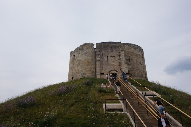 The steps up to Clifford Tower in york, with the tower at the top, with a cloudy sky, grass and people climbing the steps up to the tower