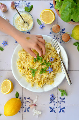 Goldgelbe Bandnudeln mit Zitronenschale und Basilikum auf einem weißen Teller. Das Teller steht auf blau-weißen Fliesen. Eine Kinderhand greift nach einem Basilikumblatt.