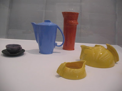 Ceramics in a museum display case. Brown cup and saucer.  Blue coffee pot.  Tall, red, cylindrical ceramic, function unclear - maybe a flower vase? Yellow  art-deco-ish, low-slung teapot and creamer.