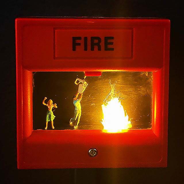 A fire alarm button with miniature people inside trying to put out a mini fire.