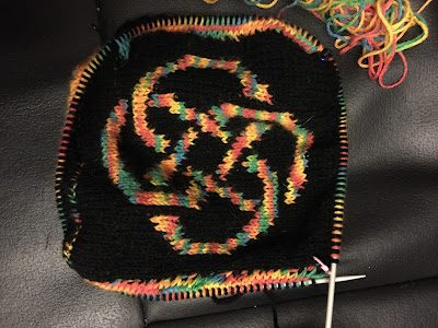 On a circular needle, a black background with a rainbow coloured Celtic knot motif. There are sections of rainbow colours just visible on two sides of the center square