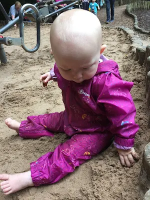 A 7 month old baby sitting in the sand in a playground and wearing a waterproof suit, but no shoes