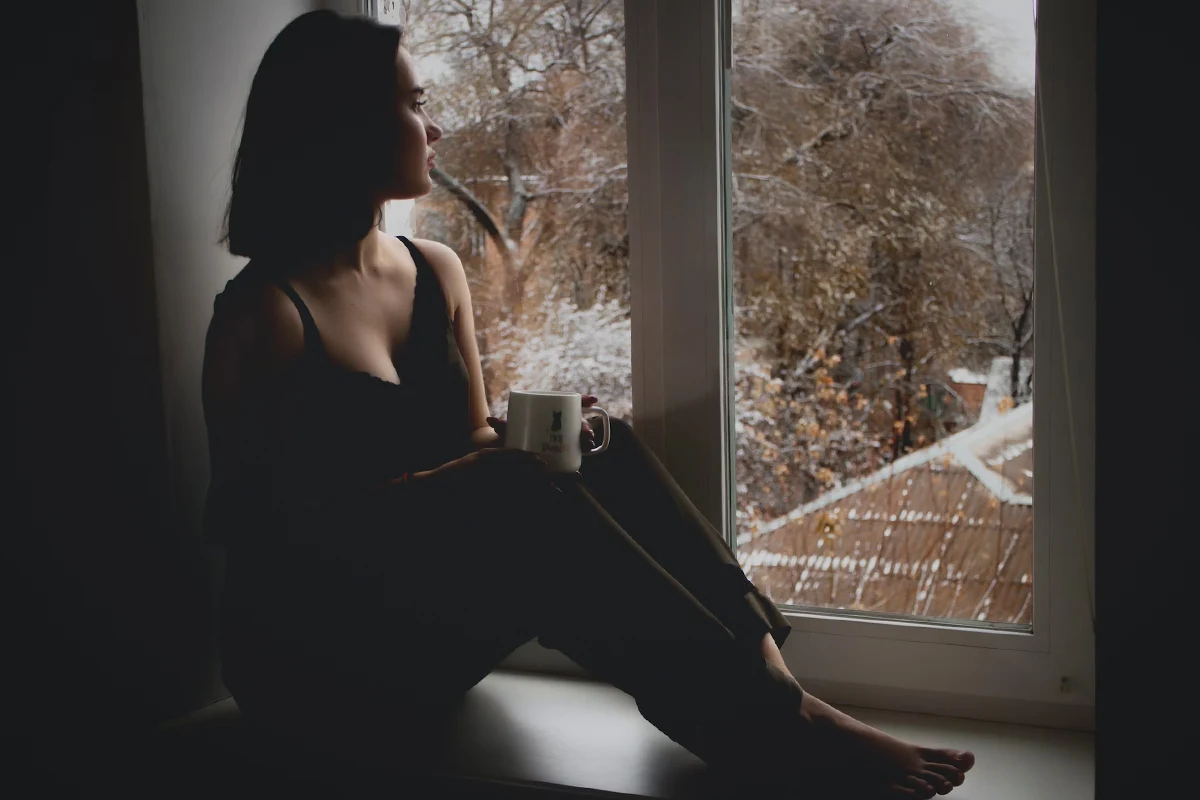 woman sitting close to the window and drinking coffee from a white mug