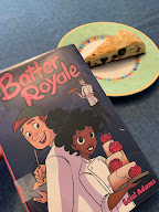 A plate with a blueberry scone on it is sitting next to the book Batter Royale. The cover has a young man and woman who are both wearing white chef coats. She is Black and is carrying a three tiered cake. He is white with red hair and is looking at her over his shoulder.